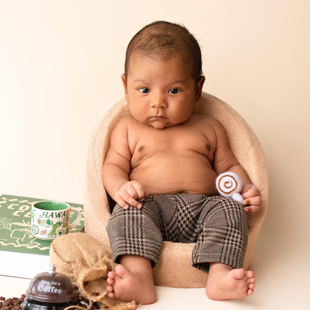 Baby in a tiny tweed armchair with houndstooth pants beside a coffee mug and felt cinnamon roll
