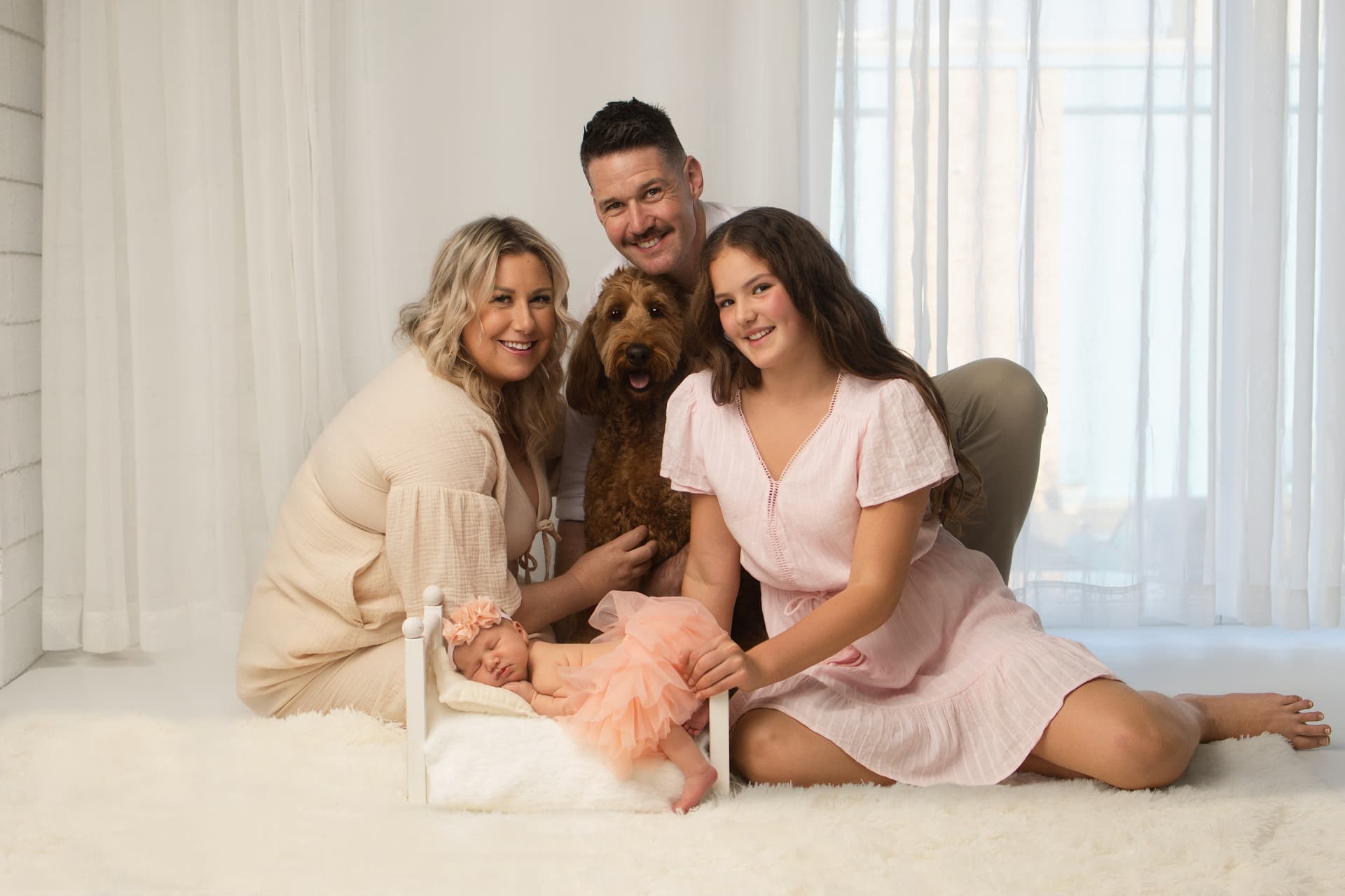 Family of four with their dog gathered around a newborn in a tiny white bed on a soft cream studio setup