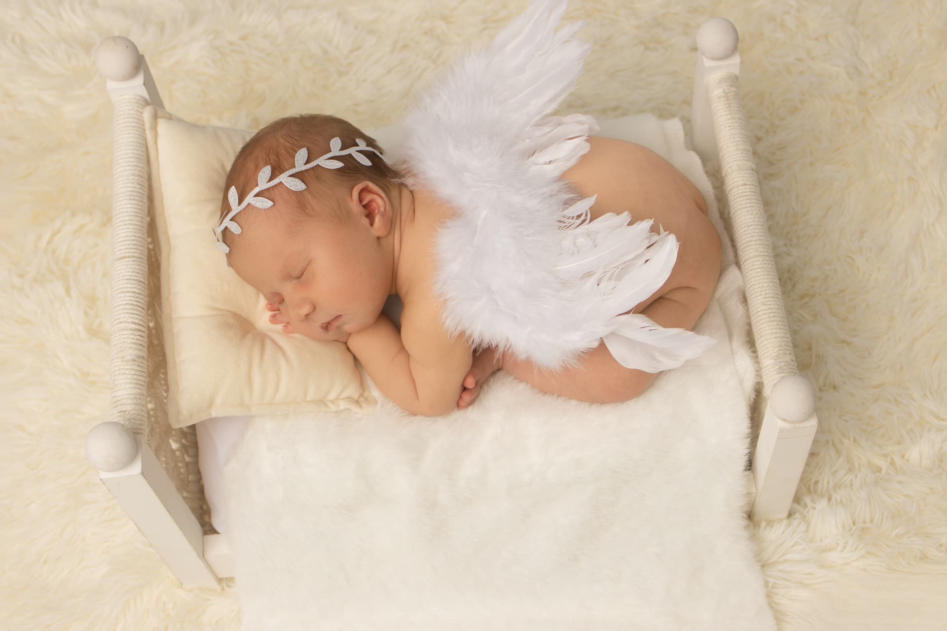 Sleeping newborn curled on a cream faux-fur rug in a miniature white wooden bed, wearing delicate white feathered angel wings and a leaf headband