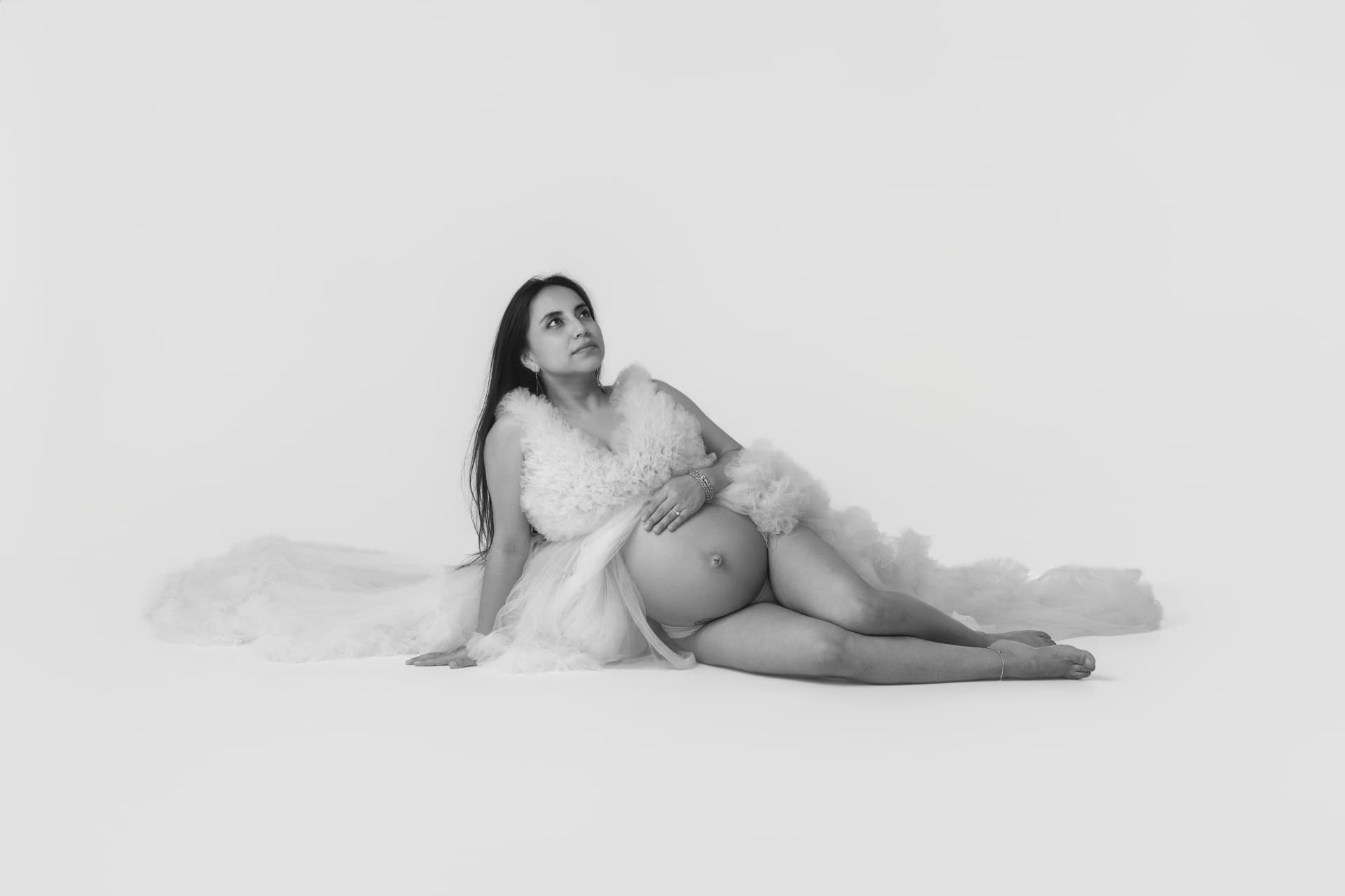 Black and white portrait of expecting mother reclining in a flowing tulle gown on a light studio backdrop, hand resting on her bump
