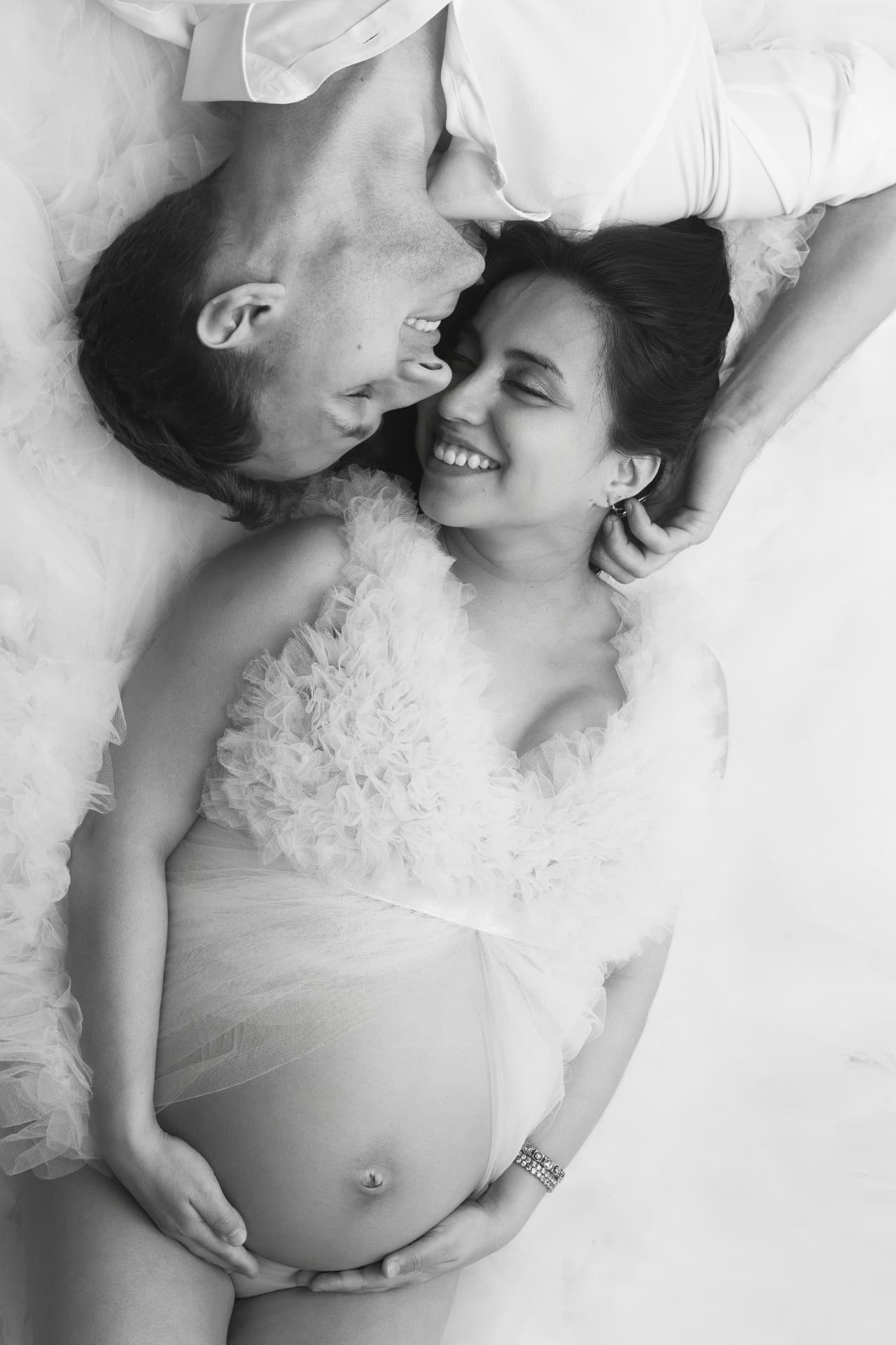 Black and white portrait of expecting couple lying on ruffled tulle, foreheads close together, hands cradling the bump