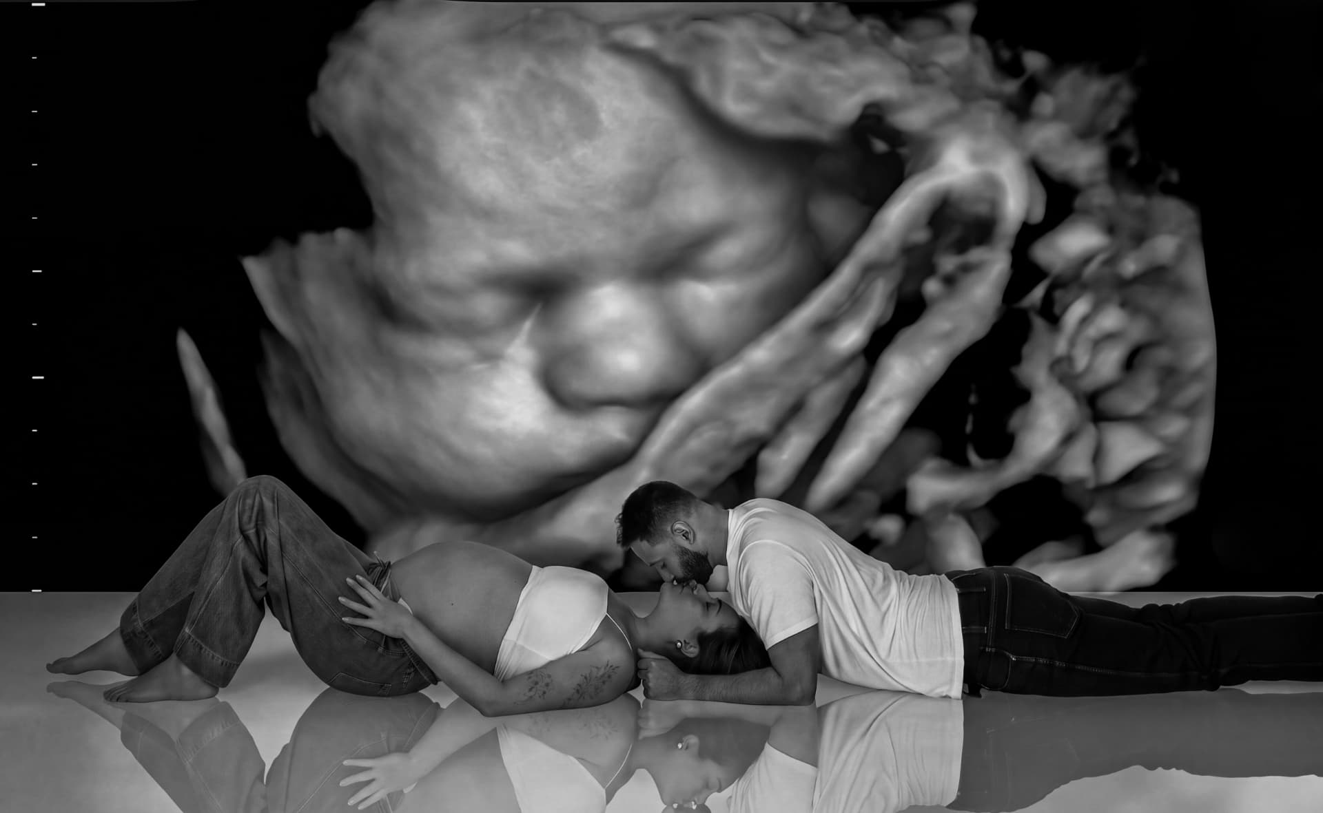 Black and white maternity portrait — an expecting couple lying close together on a reflective studio floor in front of a large ultrasound image of their baby