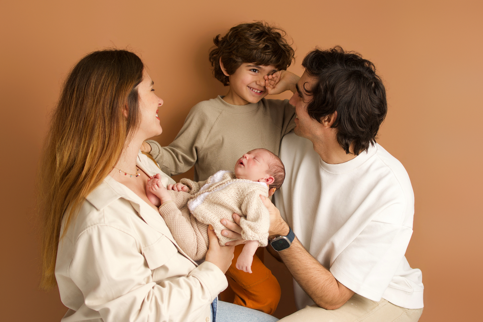 A warm family moment — parents, newborn baby and older sibling together on a soft tan backdrop