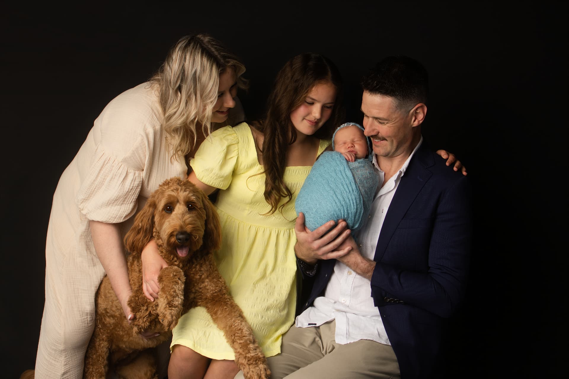 Family of five with a newborn baby and their golden curly-haired dog, gathered close against a black studio backdrop