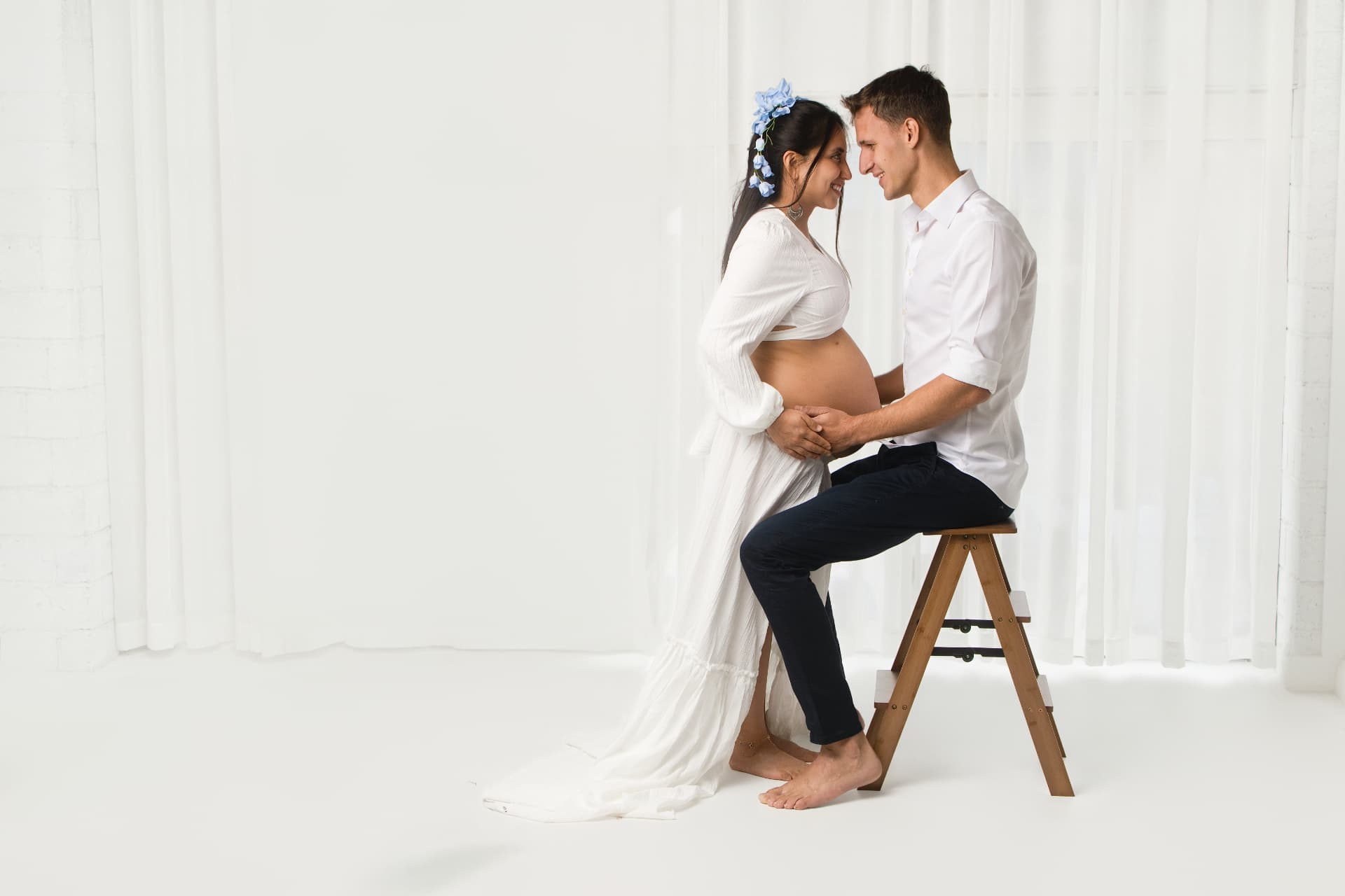 Expecting couple in white, foreheads together, hands cradling the bump beside a wooden stool