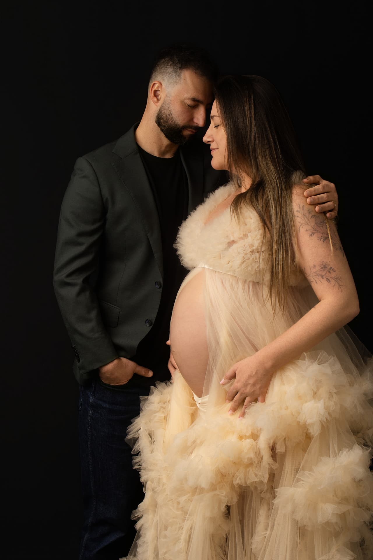 Expecting couple foreheads together on a deep black studio backdrop, mother in a cream ruffled tulle gown, partner in a dark blazer, hands cradling the bump