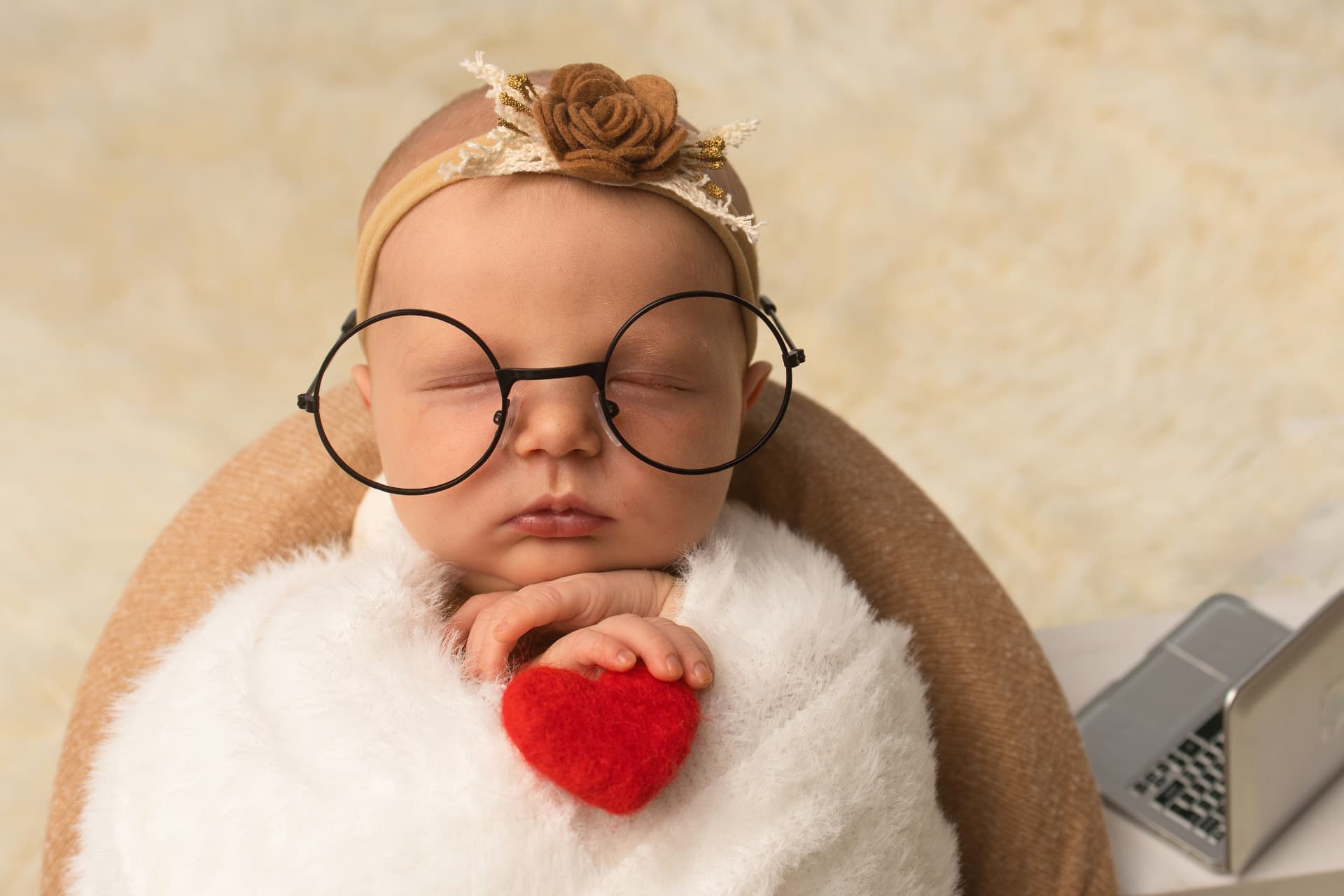 Sleeping newborn in round glasses holding a small red fuzzy heart, wrapped in a white faux-fur blanket on a cream backdrop