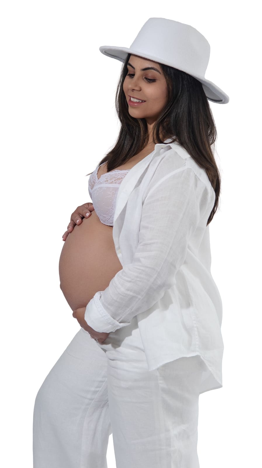Expecting mother in white linen and brimmed hat, hands resting on the bump against a clean white backdrop
