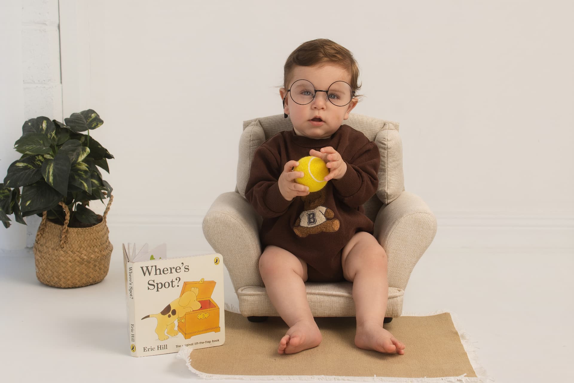 One year old in a brown teddy jumper sitting in a small armchair with glasses and a tennis ball