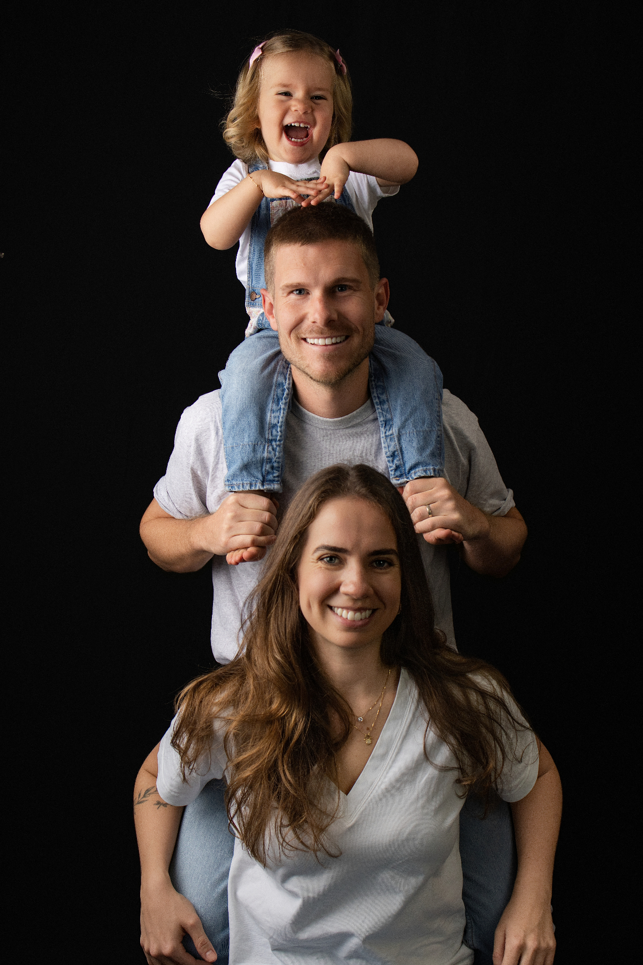 Father carrying daughter on his shoulders behind mum in a stacked family portrait on a black studio backdrop