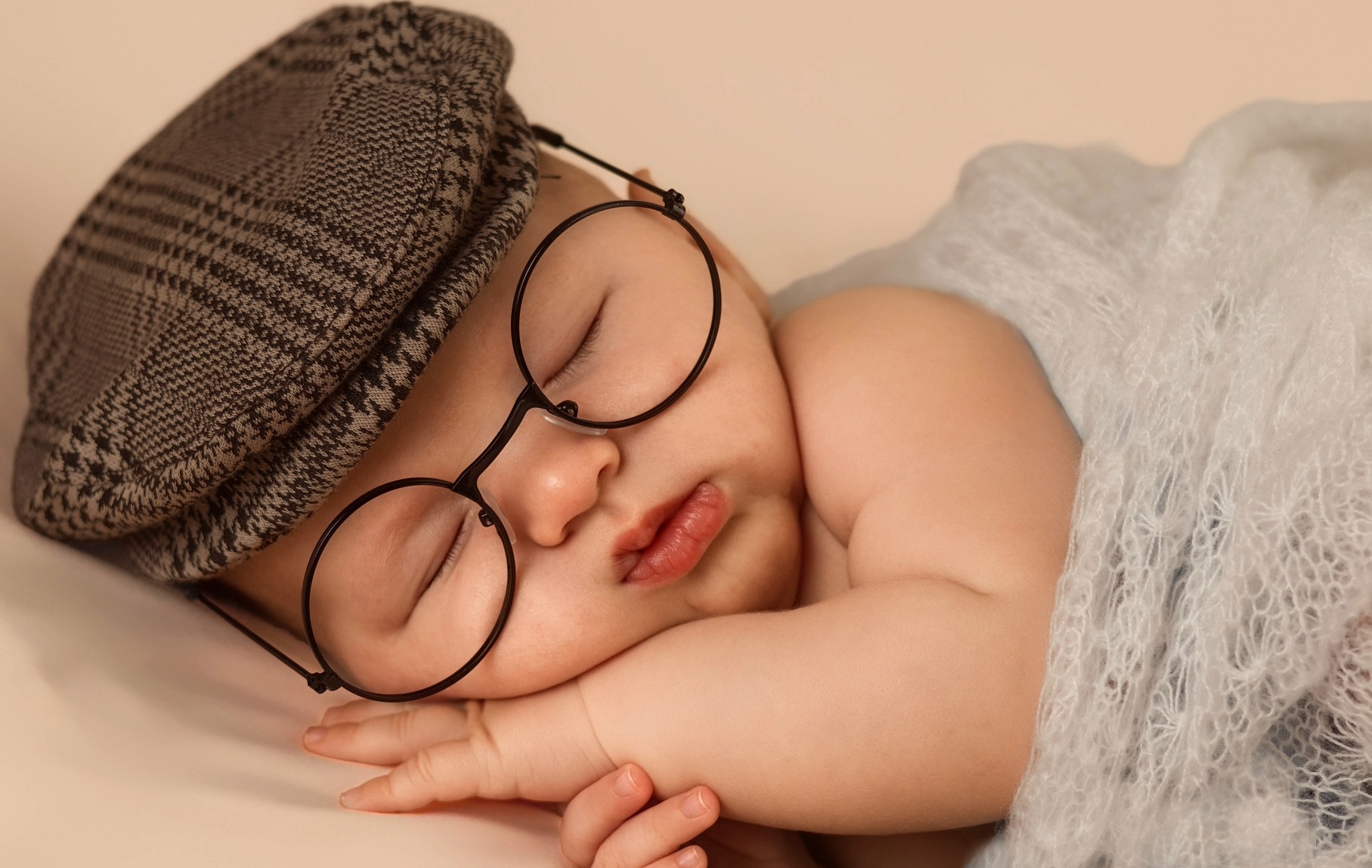Sleeping newborn in oversized round glasses and a flat cap on a soft cream backdrop