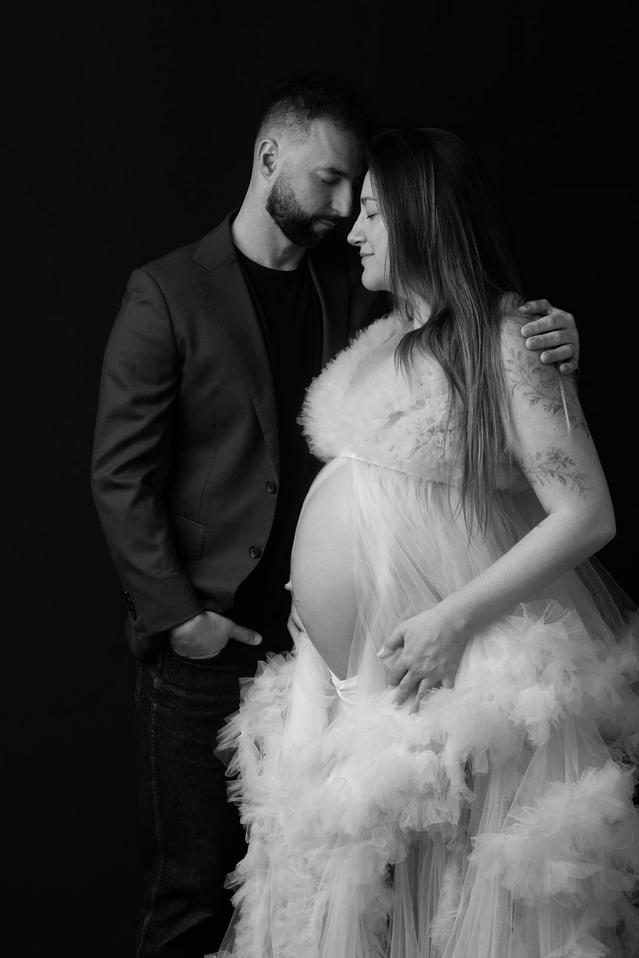 Black and white portrait of expecting couple foreheads together on a black backdrop, mother in a flowing tulle gown, hands cradling the bump
