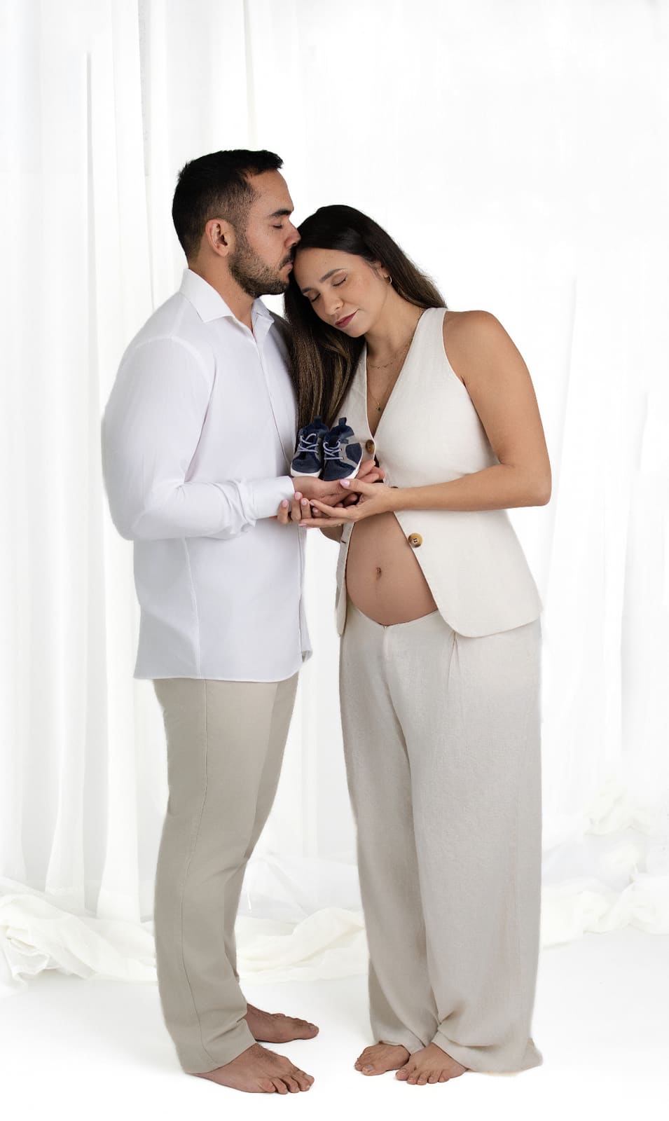 Expecting couple in soft cream wardrobe holding tiny baby shoes against the bump in front of sheer white curtains