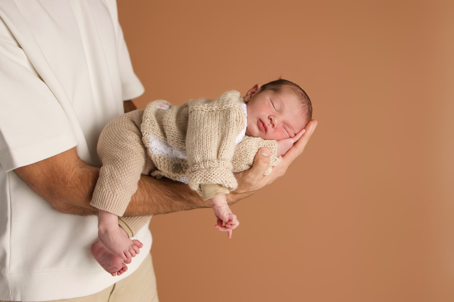 Father holding a sleeping newborn in a knitted outfit against a warm brown studio backdrop