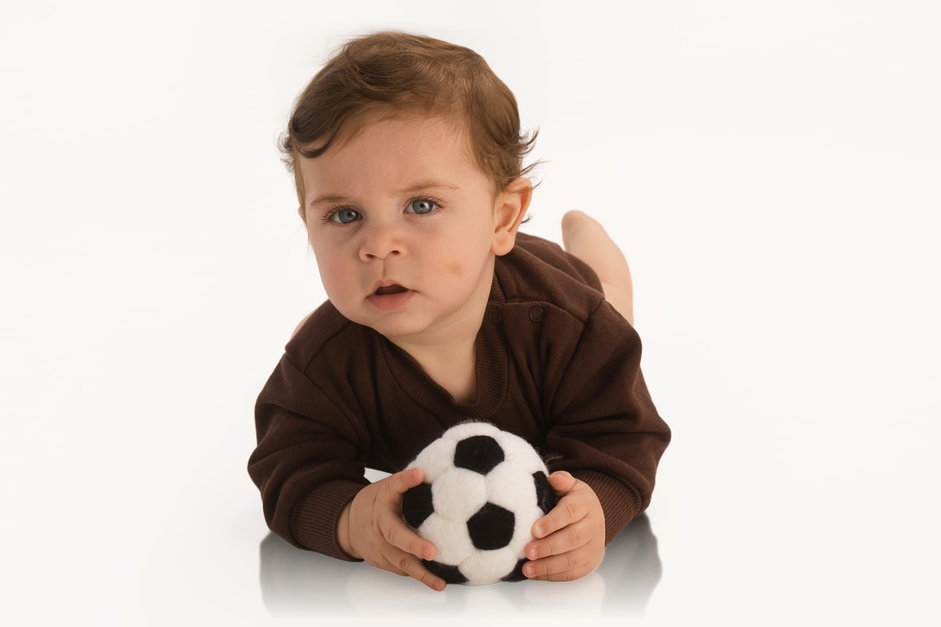 Close-up of a baby lying on a bright white surface, propped on elbows and holding a small black-and-white plush soccer ball