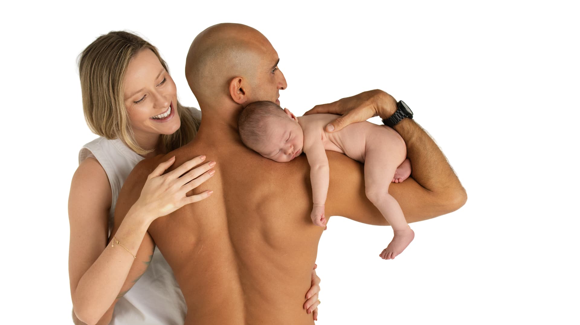 Newborn resting on father's shoulder while mother smiles beside them on a white studio backdrop