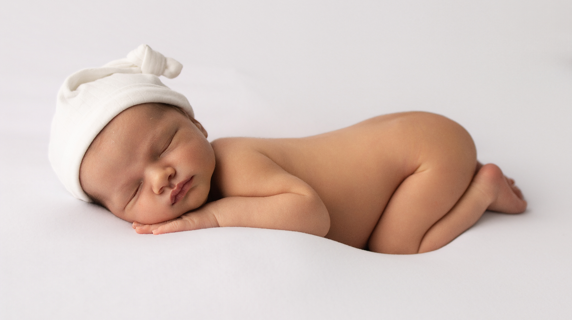 Sleeping newborn in a soft cream knot hat, curled on a smooth cream backdrop